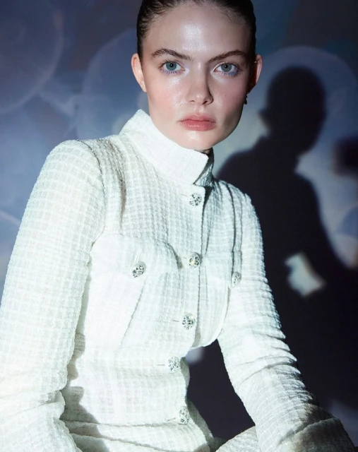 Close-up studio portrait of a young woman with fair skin, wearing a light-colored jacket with decorative buttons, against a blurred background.