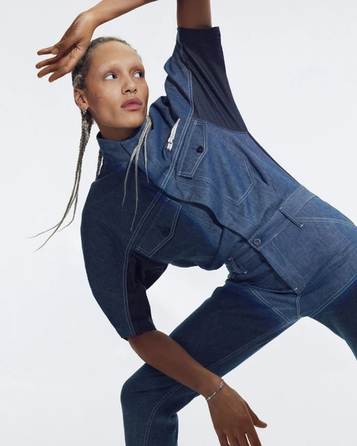 A fair-skinned adult woman in a blue denim jumpsuit poses against a white backdrop, showcasing a fashion editorial style with braided hair.