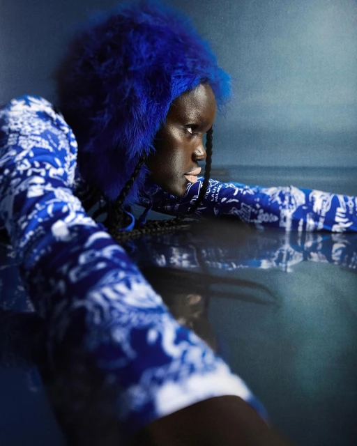 A woman with dark skin and blue faux fur head covering, wearing a patterned blue top, poses in a close up shot with dark water.