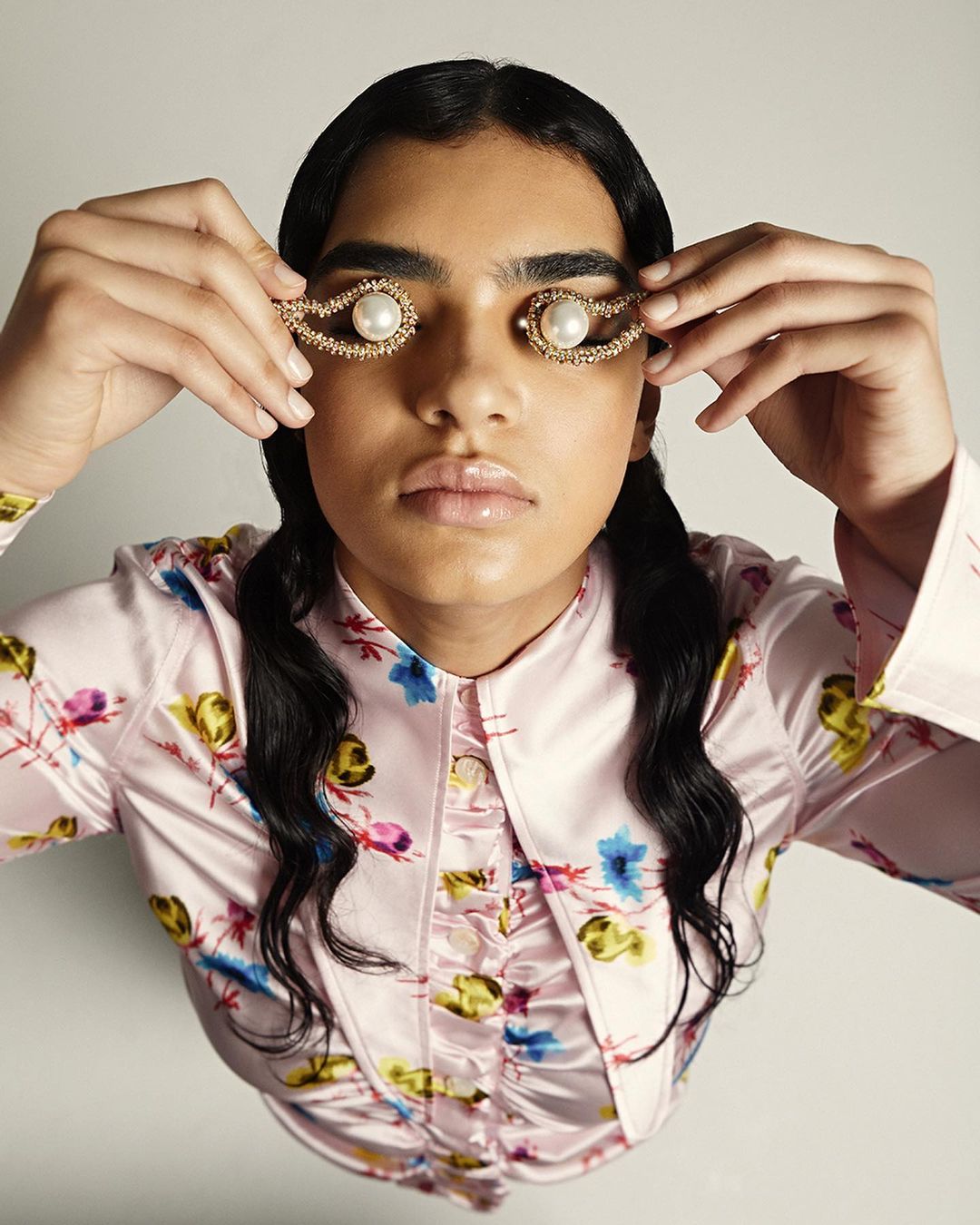 Woman with dark hair wears a floral top and eye jewelry with pearls, looking up at the camera in an indoor setting against a white background.