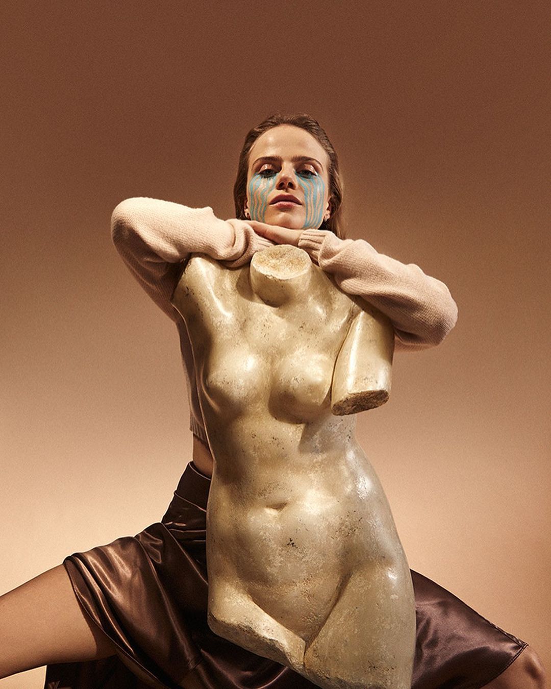Woman with blue face paint poses with a white torso sculpture against a tan backdrop, wearing a pink top and brown skirt.