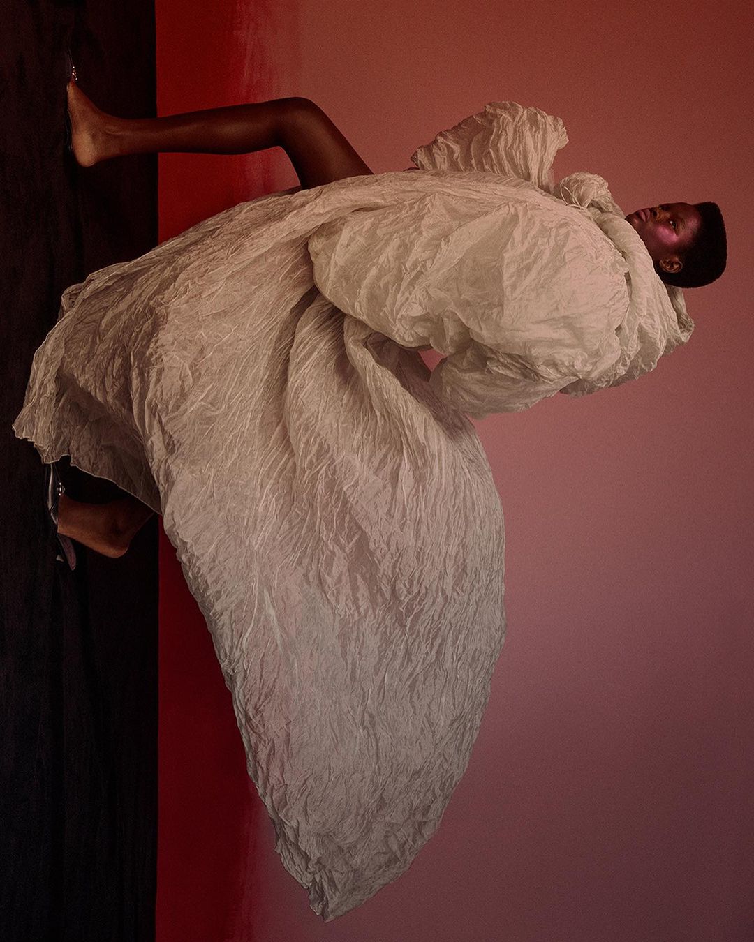 Woman in large textured white coat poses against gradient pink and black background in fashion editorial-style studio shot.