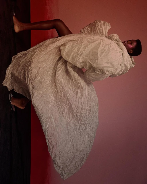 Woman in large textured white coat poses against gradient pink and black background in fashion editorial-style studio shot.