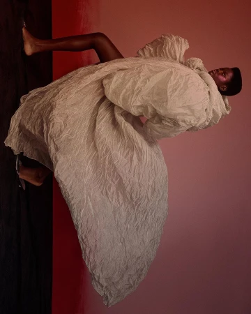 Woman in large textured white coat poses against gradient pink and black background in fashion editorial-style studio shot.