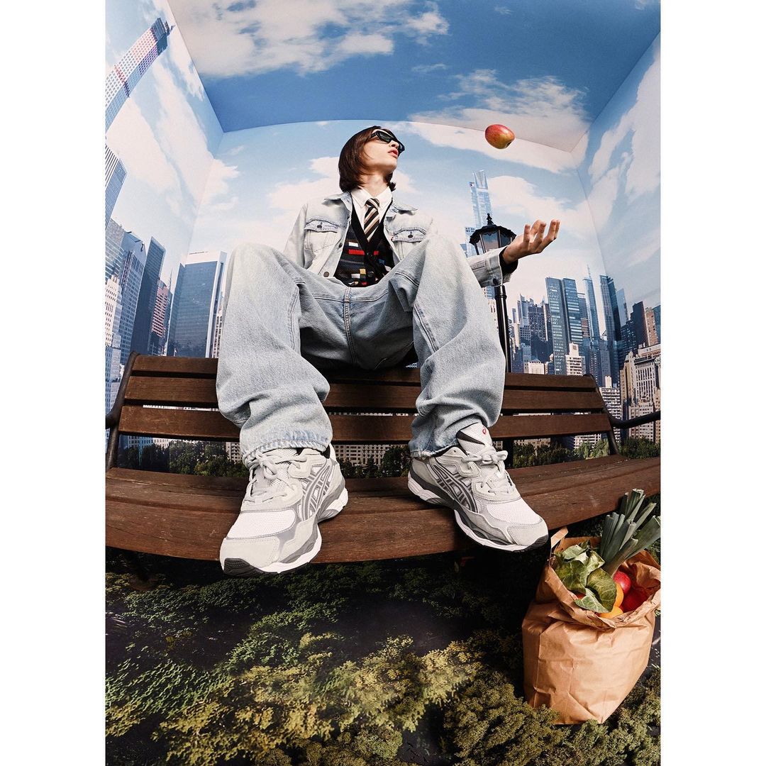 A person in sunglasses sits on a wooden bench indoors with a cityscape mural backdrop, an apple floating above their hand.