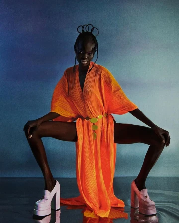 Black model in bright orange dress and platform loafers squatting against blue gradient background by Katia Wik