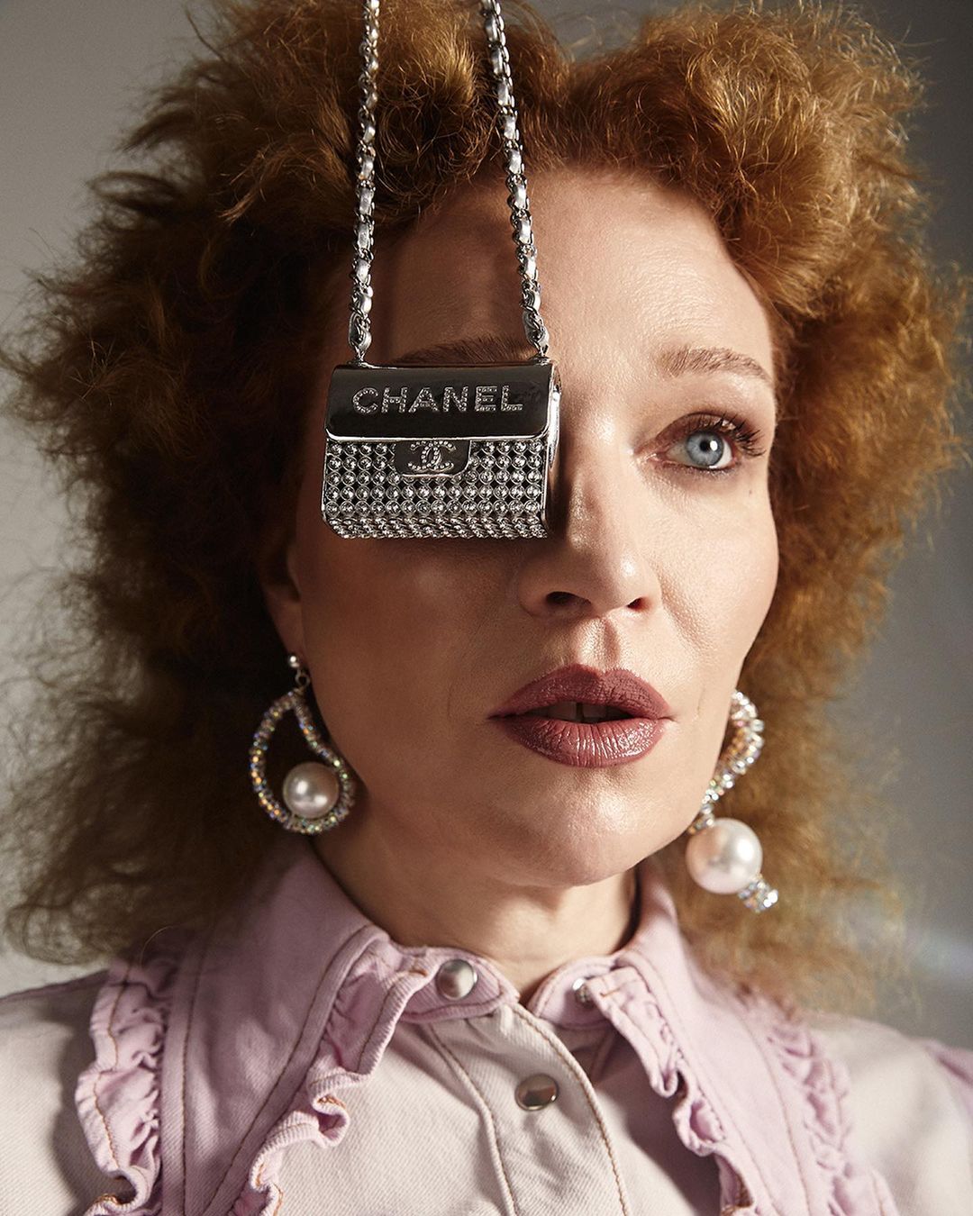 Woman with red hair poses with a Chanel purse covering one eye, accessorized with pearl earrings and a ruffled pink shirt.
