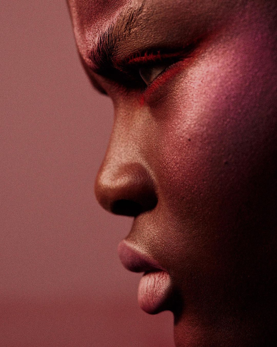 Close-up studio portrait of a Black woman with red eye makeup, pink skin tones, and a neutral expression in front of a pink backdrop.