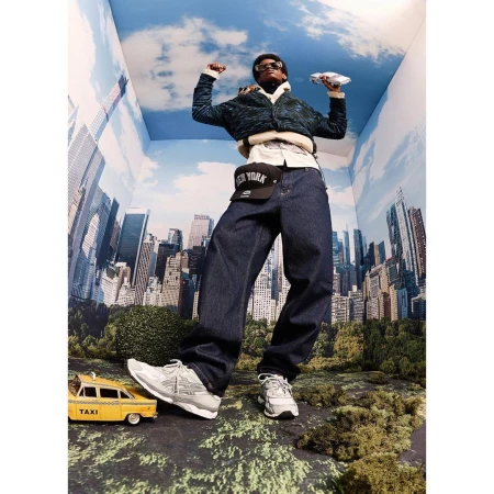 A man in dark denim and sneakers poses in a photo studio surrounded by a NYC skyscape backdrop, greenery, and a toy taxi.