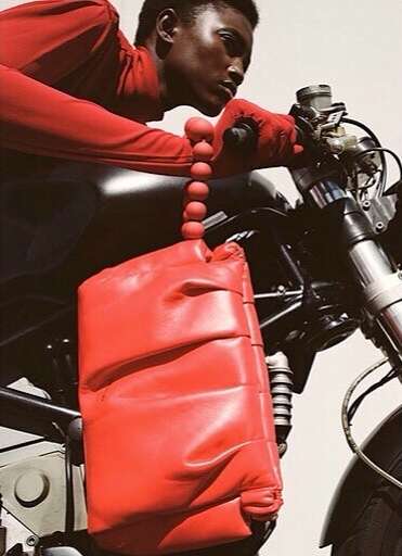 An African-American adult male in red clothing sits on a black motorcycle, holding a large red padded shoulder bag, creating a bold fashion statement.