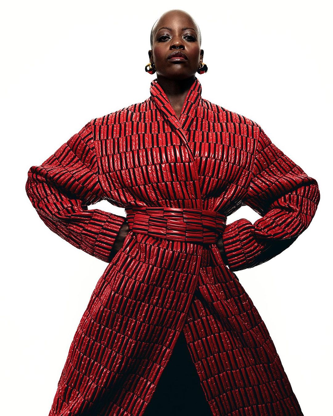 Portrait of Black adult female wearing a striking red textured coat dress, posing with hands on hips against a stark white background.