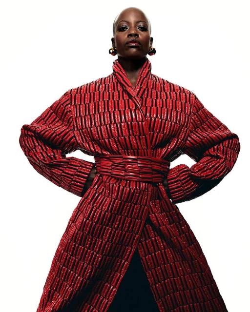 Portrait of Black adult female wearing a striking red textured coat dress, posing with hands on hips against a stark white background.