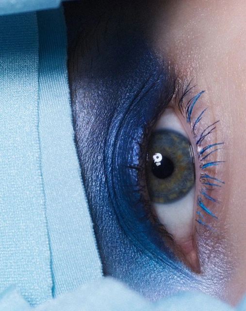 Close-up of a woman's eye with vibrant blue makeup on the eyelid and lashes, set against a light blue soft fabric.