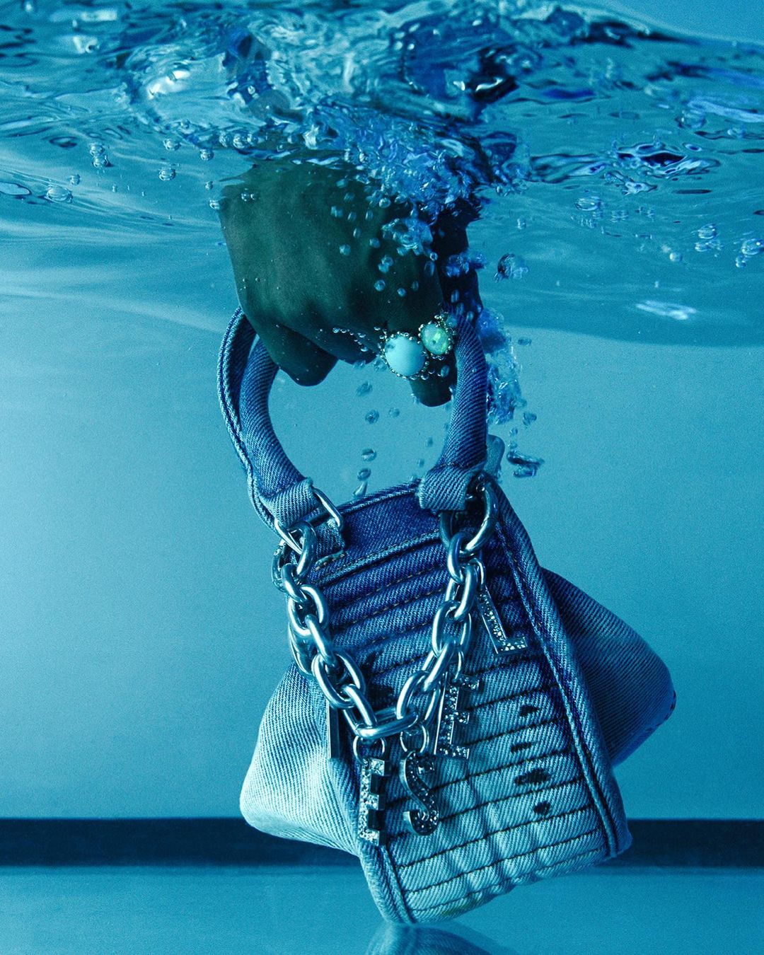 Denim purse with chain and charms held by a gloved hand underwater with blue lighting and visible water bubbles near the surface.