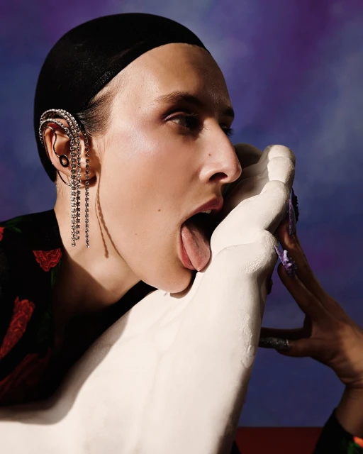 Fashion image of model with long earring licking white sculpture on a blue background, floral black top, long purple nails, studio lighting.