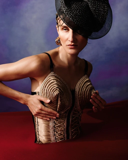 Woman in elegant gold corset and black hat poses against blurred purple background. Fashion portrait with artistic costume and dramatic lighting.