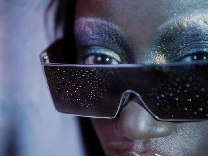 Close-up beauty shot of an adult woman with futuristic sunglasses and silver makeup. The background has an artistic, airbrushed look.