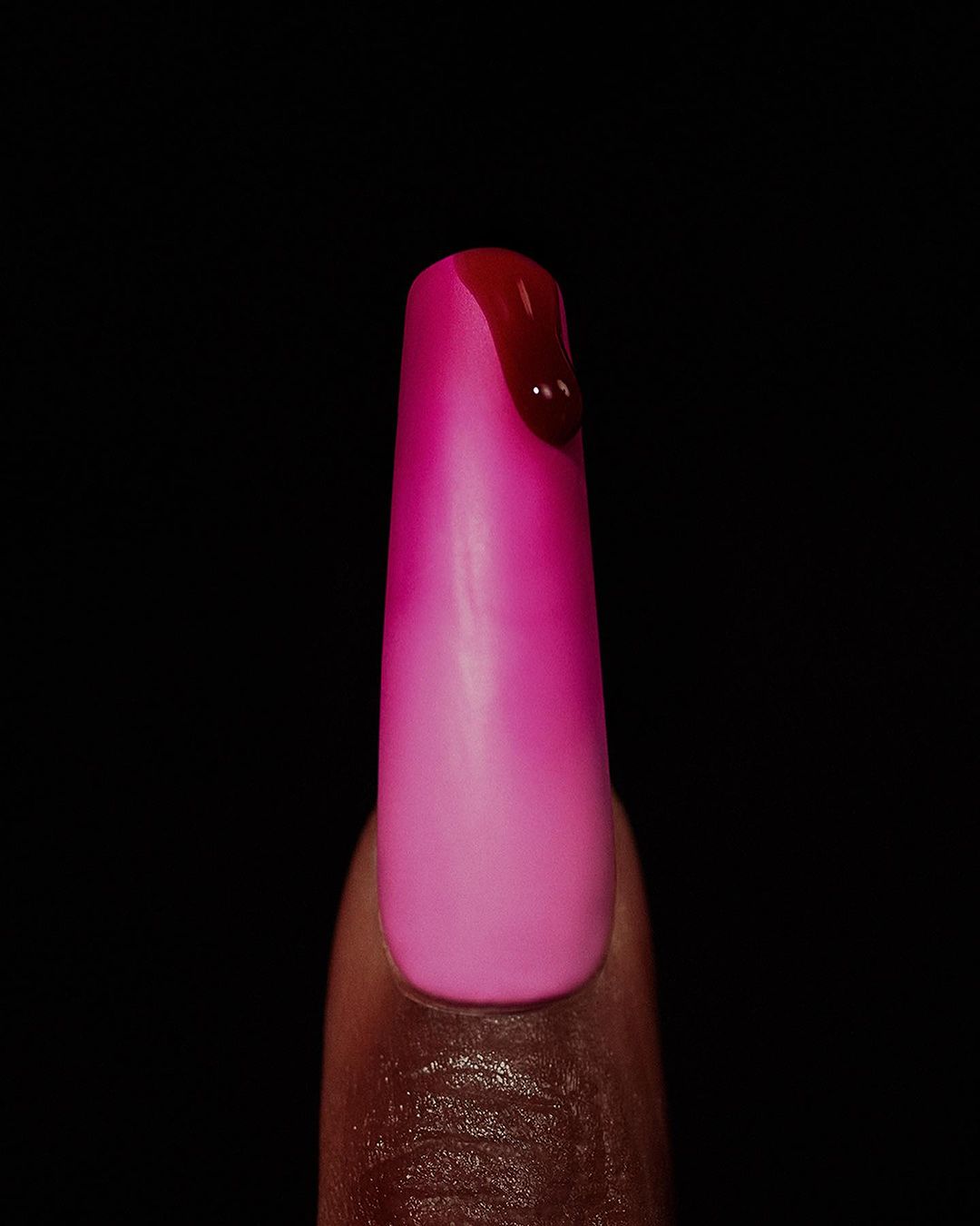 Close-up of a manicured fingernail with pink and magenta ombre nail polish and a drip of red liquid on a dark black background.