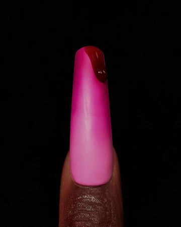 Close-up of a manicured fingernail with pink and magenta ombre nail polish and a drip of red liquid on a dark black background.