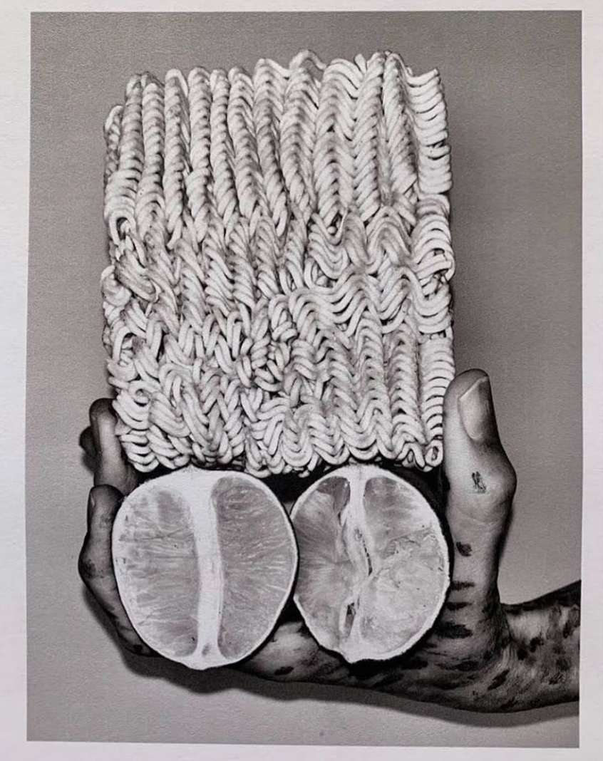 Monochrome photograph of a block of ramen noodles above two halved oranges, held in a hand, showcasing texture and abstract food presentation.
