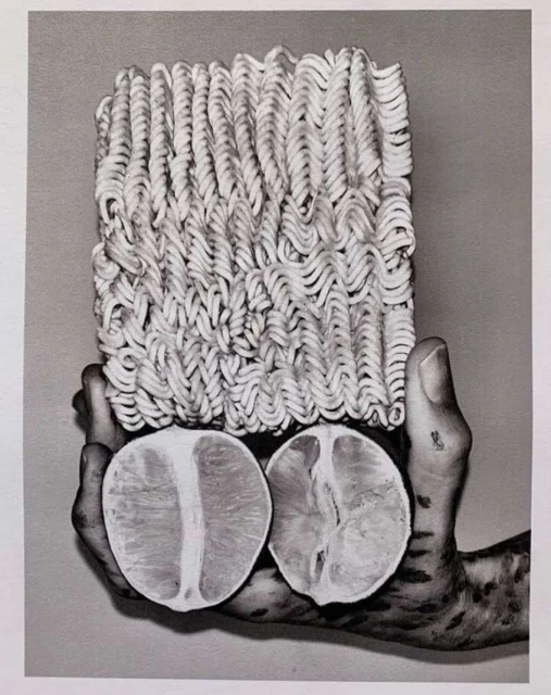 Monochrome photograph of a block of ramen noodles above two halved oranges, held in a hand, showcasing texture and abstract food presentation.