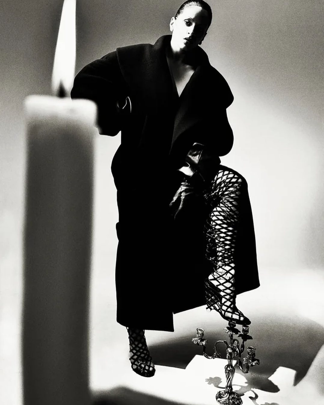 Monochrome fashion shot of a woman in a long coat and fishnet stockings, stepping on a candleholder next to a tall candle.