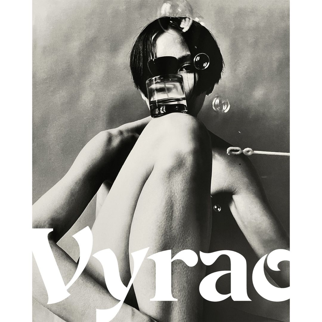Artistic black and white shot of a person with a short dark haircut, partially nude, holding a perfume bottle and surrounded by bubbles.