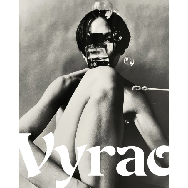 Artistic black and white shot of a person with a short dark haircut, partially nude, holding a perfume bottle and surrounded by bubbles.