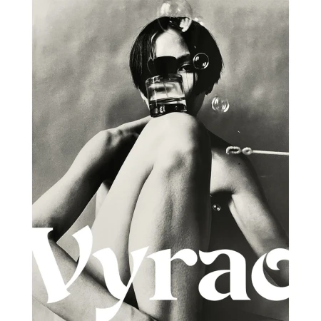 Artistic black and white shot of a person with a short dark haircut, partially nude, holding a perfume bottle and surrounded by bubbles.