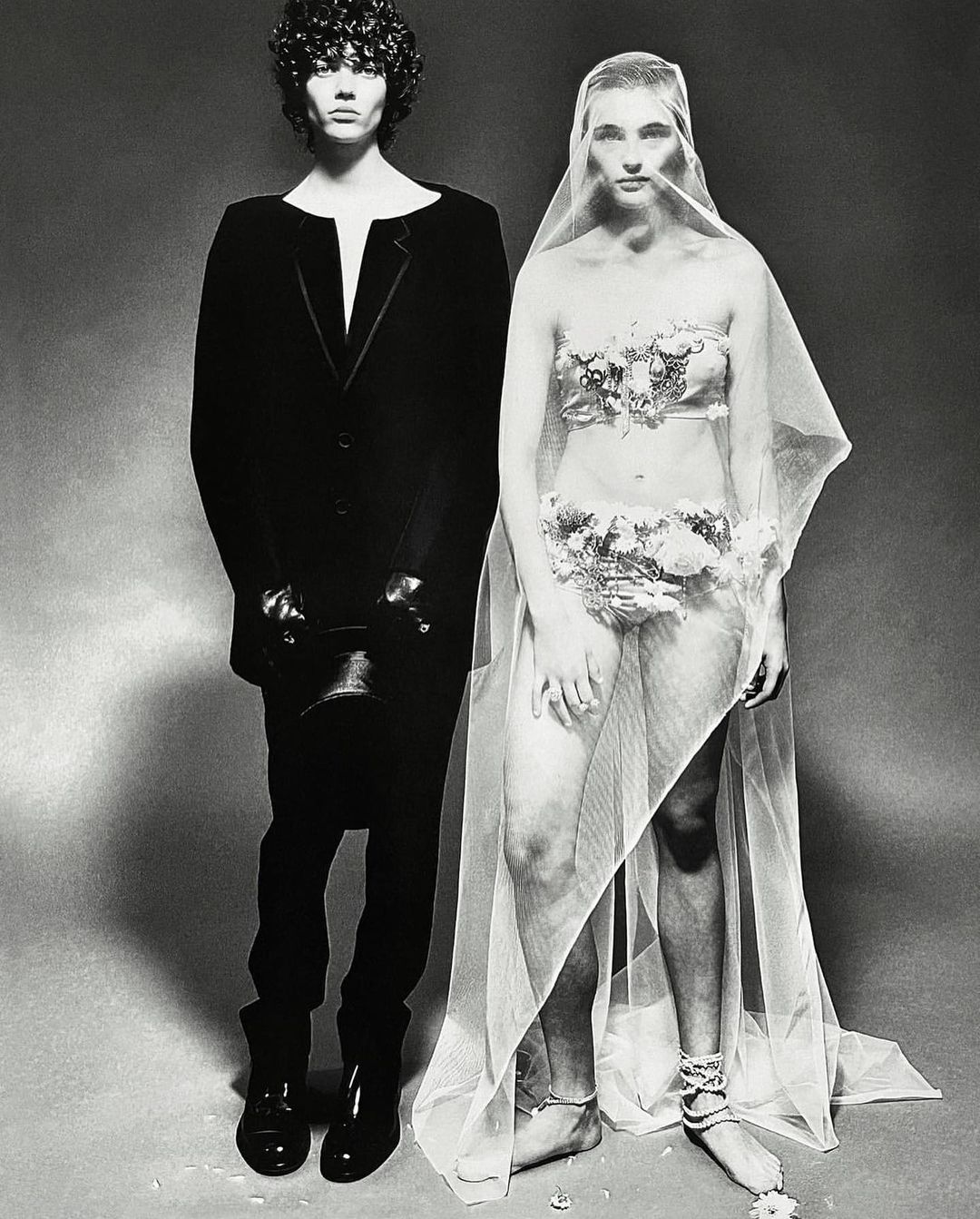 Monochrome studio shot of a man in a black suit and a woman with floral accents, partially veiled, set against a neutral backdrop.