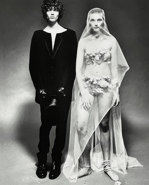 Monochrome studio shot of a man in a black suit and a woman with floral accents, partially veiled, set against a neutral backdrop.