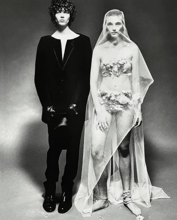 Monochrome studio shot of a man in a black suit and a woman with floral accents, partially veiled, set against a neutral backdrop.