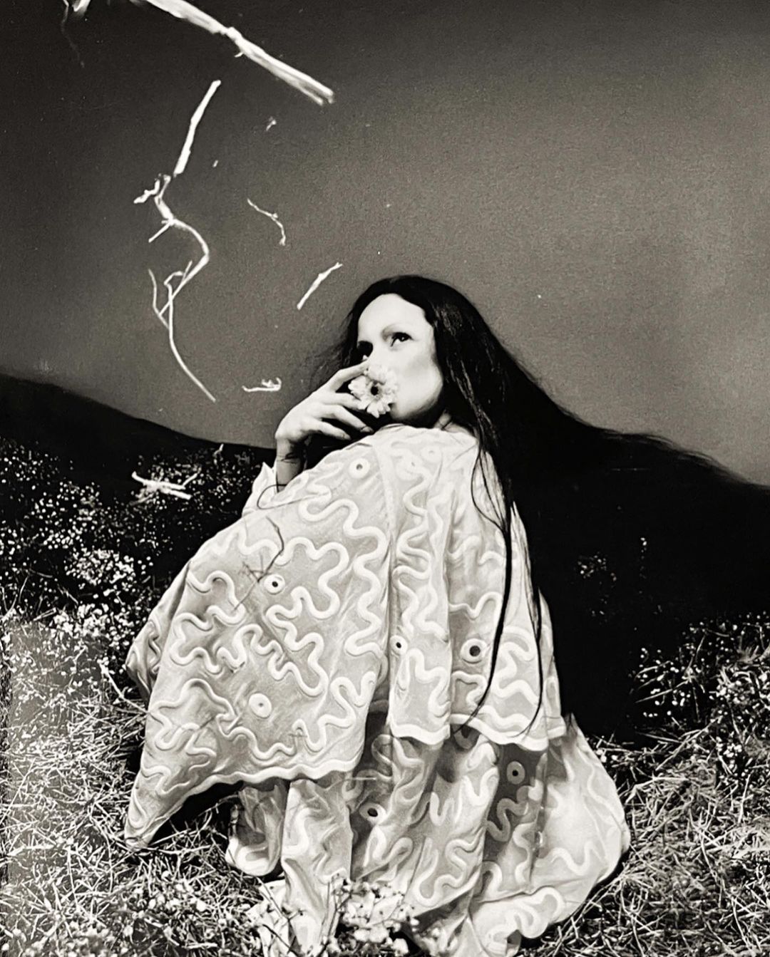 Black and white portrait of a woman in a patterned shawl, seated in a field, with branches in the background, exuding a fine art style.