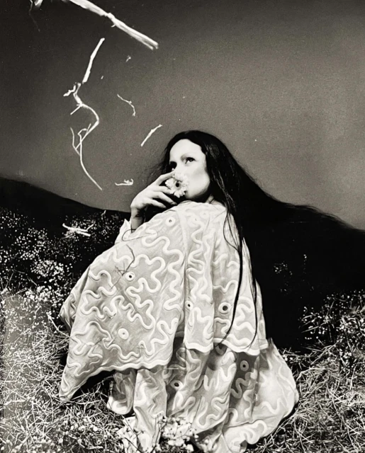 Black and white portrait of a woman in a patterned shawl, seated in a field, with branches in the background, exuding a fine art style.