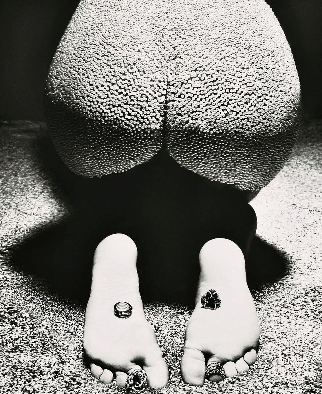 High-contrast studio shot in black and white: a person kneeling with textured skin, multiple rings on toes and fingers, and dark background.