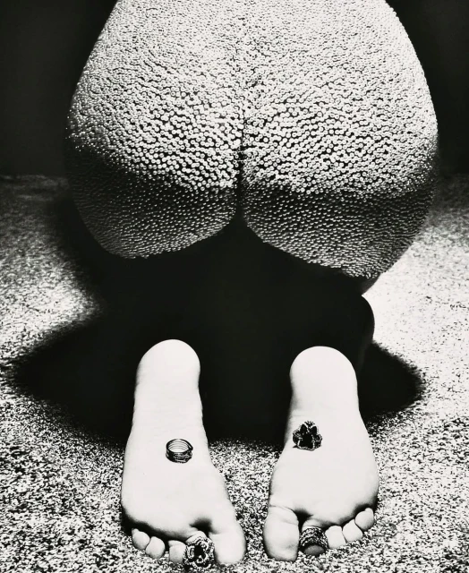 High-contrast studio shot in black and white: a person kneeling with textured skin, multiple rings on toes and fingers, and dark background.