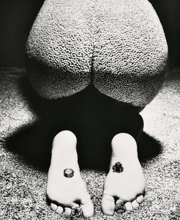 High-contrast studio shot in black and white: a person kneeling with textured skin, multiple rings on toes and fingers, and dark background.