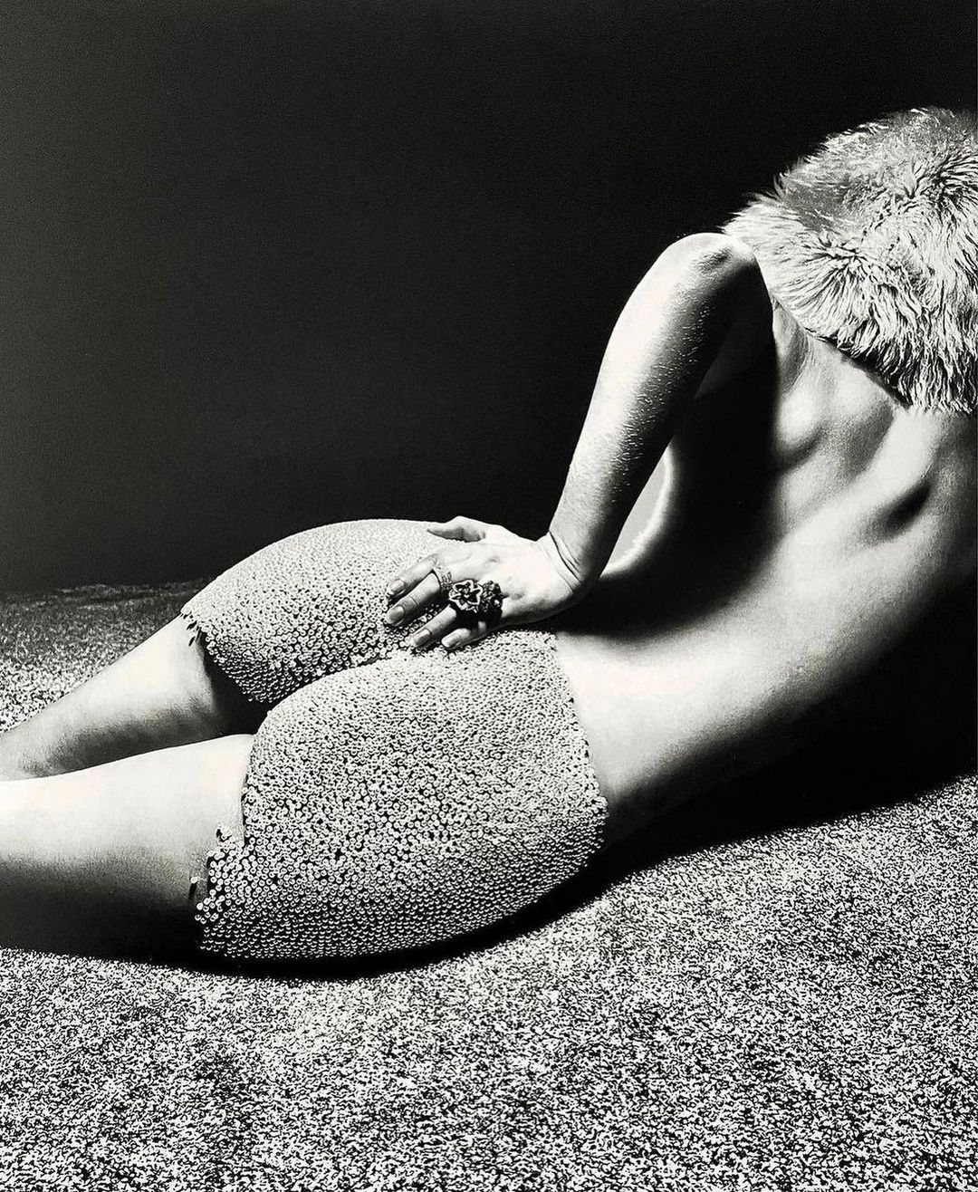 Monochrome artistic photograph of a semi-nude woman lying on textured surface, wearing decorative shorts and fur collar.