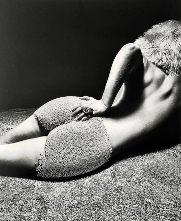 Monochrome artistic photograph of a semi-nude woman lying on textured surface, wearing decorative shorts and fur collar.