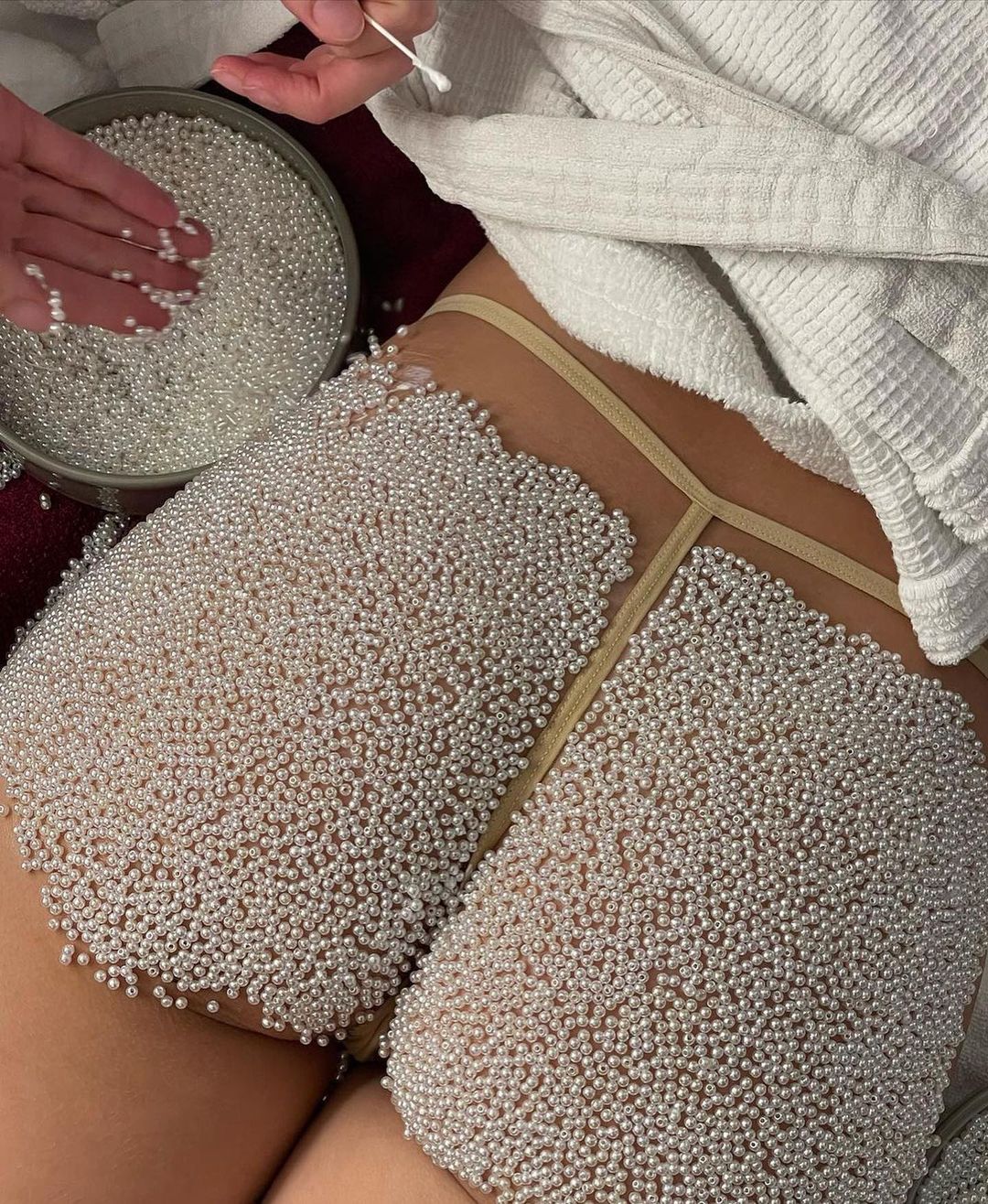 Close-up of a person's thighs and torso covered in small white pearls, hands placing pearls from a container, white robe in background.