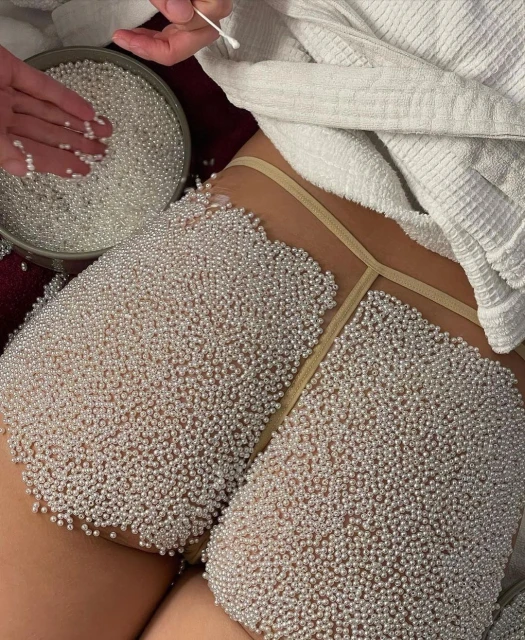Close-up of a person's thighs and torso covered in small white pearls, hands placing pearls from a container, white robe in background.