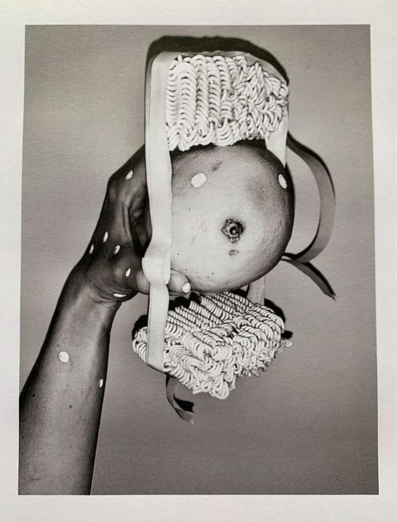 A black and white studio shot presents an arm and hand holding a fruit with noodle-covered hair accessories against a neutral backdrop.