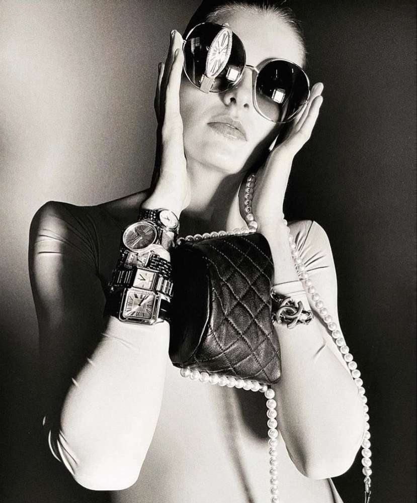 A woman with oversized sunglasses, multiple watches, and a pearl necklace poses in a black-and-white fashion shot.