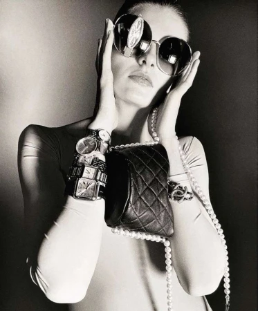 A woman with oversized sunglasses, multiple watches, and a pearl necklace poses in a black-and-white fashion shot.