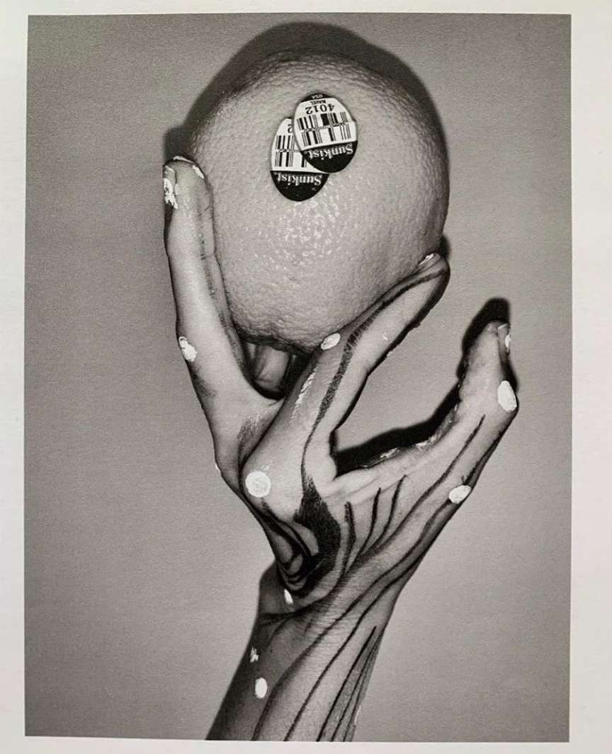 Monochrome art photograph of a hand with painted linear details holding a Sunkist orange, captured in a studio setting with soft lighting.