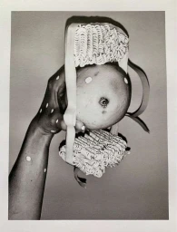 Hand holding a citrus fruit adorned with instant ramen noodles and ribbon in a conceptual black and white photograph.