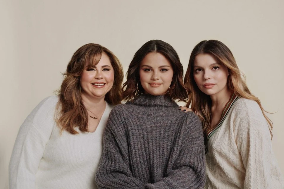 Three women pose indoors. Selena Gomez, in the middle, wears a grey sweater. Flanking her are two women in light colored sweaters.