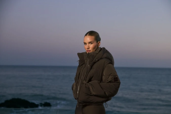 Adult woman in a puffer coat stands near the ocean against a dusky sky in a medium shot with a shallow depth of field, outdoors.