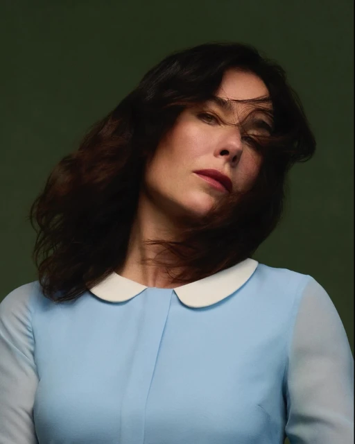 Portrait of a brunette woman with fair skin and a blue dress with a white collar against a dark green background in a studio setting.