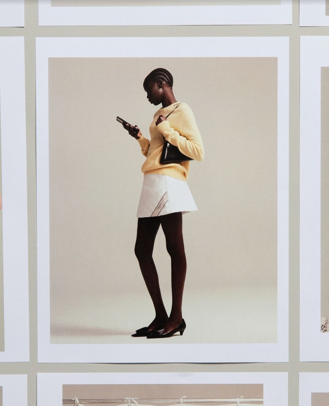 Black woman in a yellow sweater and white skirt holding a smartphone against a pale background